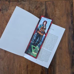 Puede incluir: Un marcapáginas de vidrio emplomado con una mujer vestida con un atuendo azul y marrón de pie sobre un fondo verde y marrón. El marcapáginas tiene un borde rojo y el texto "Capítulo Cuatro" en la esquina inferior derecha.