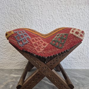 May include: A small, folding wooden stool with a woven seat. The seat has a red background with geometric patterns in blue, green, and white. The wooden legs are intricately carved with decorative patterns.