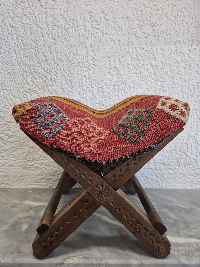 May include: A small, folding wooden stool with a woven seat. The seat has a red background with geometric patterns in blue, green, and white. The wooden legs are intricately carved with decorative patterns.