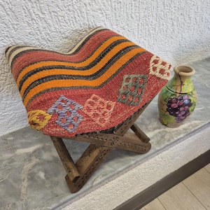 May include: A wooden stool with a woven seat. The seat has a red, orange, and black striped pattern with embroidered geometric designs. A decorative vase with a grape design is also visible.