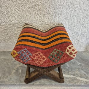 May include: A small wooden stool with a woven seat. The seat has a pattern of red, orange, and black stripes, and diamond-shaped designs in various colours. The wooden legs are intricately carved. The stool is approximately 30 cm in height.
