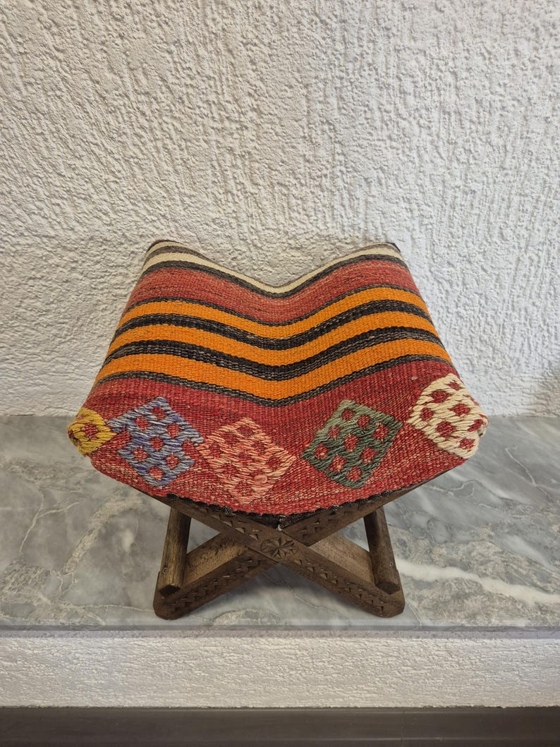 May include: A small wooden stool with a woven seat. The seat has a pattern of red, orange, and black stripes, and diamond-shaped designs in various colours. The wooden legs are intricately carved. The stool is approximately 30 cm in height.