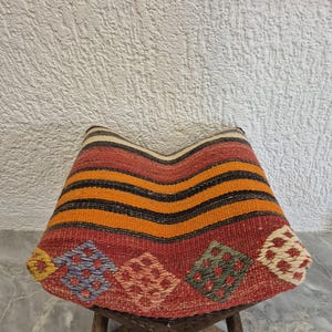 May include: A handcrafted, colourful, woven stool with a patterned seat. The seat features horizontal stripes in orange and black, and diamond-shaped patterns in red, blue, and green. The wooden legs are intricately carved.