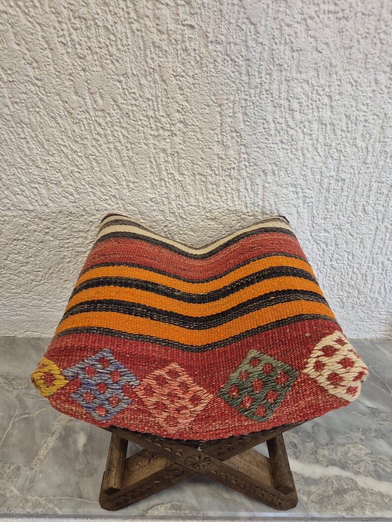 May include: A handcrafted, colourful, woven stool with a patterned seat. The seat features horizontal stripes in orange and black, and diamond-shaped patterns in red, blue, and green. The wooden legs are intricately carved.