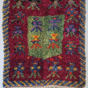 May include: A rectangular, hand-woven rug with a deep red central field and a green central square. The rug features colorful floral and geometric patterns in blue, orange, yellow, and purple. The border is a mix of blue and yellow zigzags. The rug has a fringe.