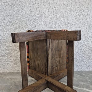 May include: A wooden stool with a woven fabric seat. The stool has a dark brown finish and a cross-legged base. The seat features a colourful pattern with orange, red, and brown hues. The stool is sturdy and appears to be handmade.