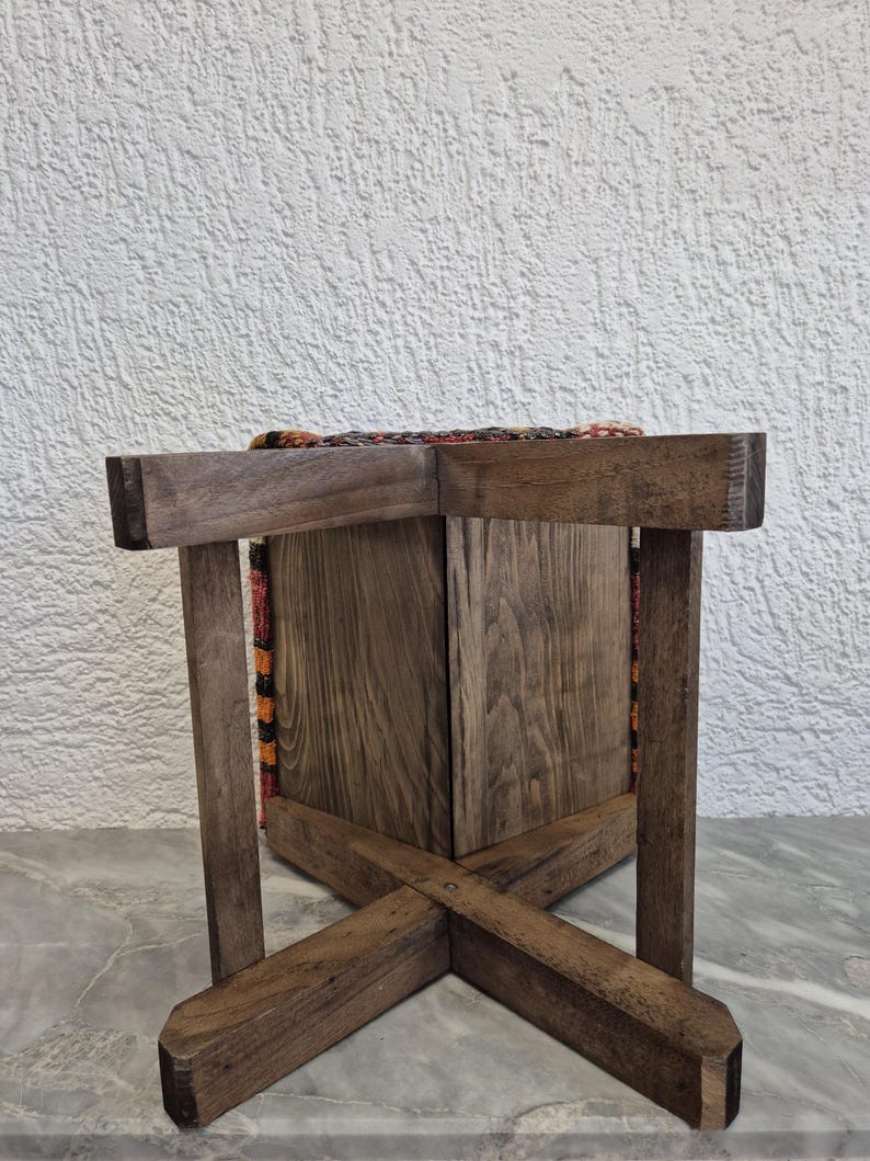 May include: A wooden stool with a woven fabric seat. The stool has a dark brown finish and a cross-legged base. The seat features a colourful pattern with orange, red, and brown hues. The stool is sturdy and appears to be handmade.