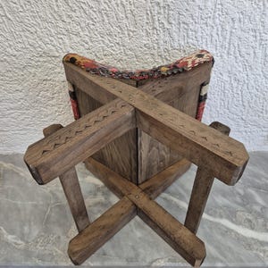 May include: A wooden stool with a cross-legged base and a patterned fabric seat. The wood has a natural finish with carved details. The seat features a colourful, geometric design with red, yellow, and black accents. The stool is designed for sitting.