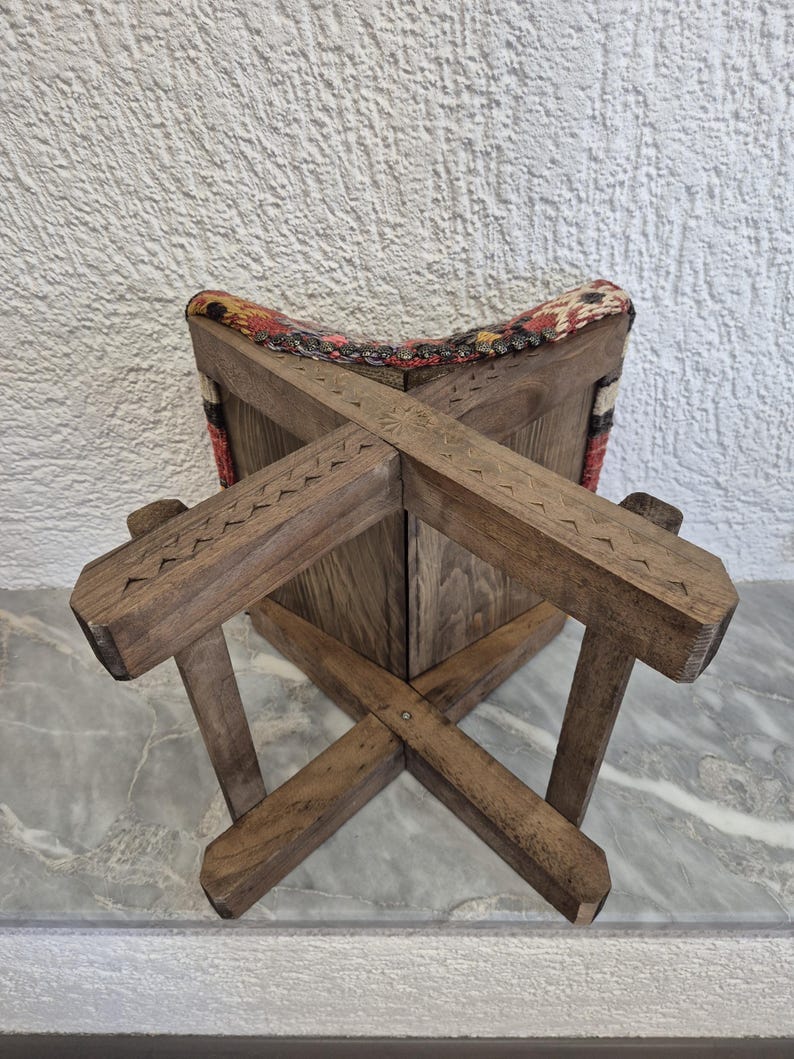 May include: A wooden stool with a cross-legged base and a patterned fabric seat. The wood has a natural finish with carved details. The seat features a colourful, geometric design with red, yellow, and black accents. The stool is designed for sitting.