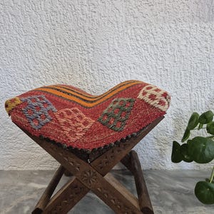 May include: A small, folding wooden stool with a patterned seat. The seat has a red background with colourful geometric designs. The wooden frame has carved details. The stool is designed for sitting and is a decorative item.