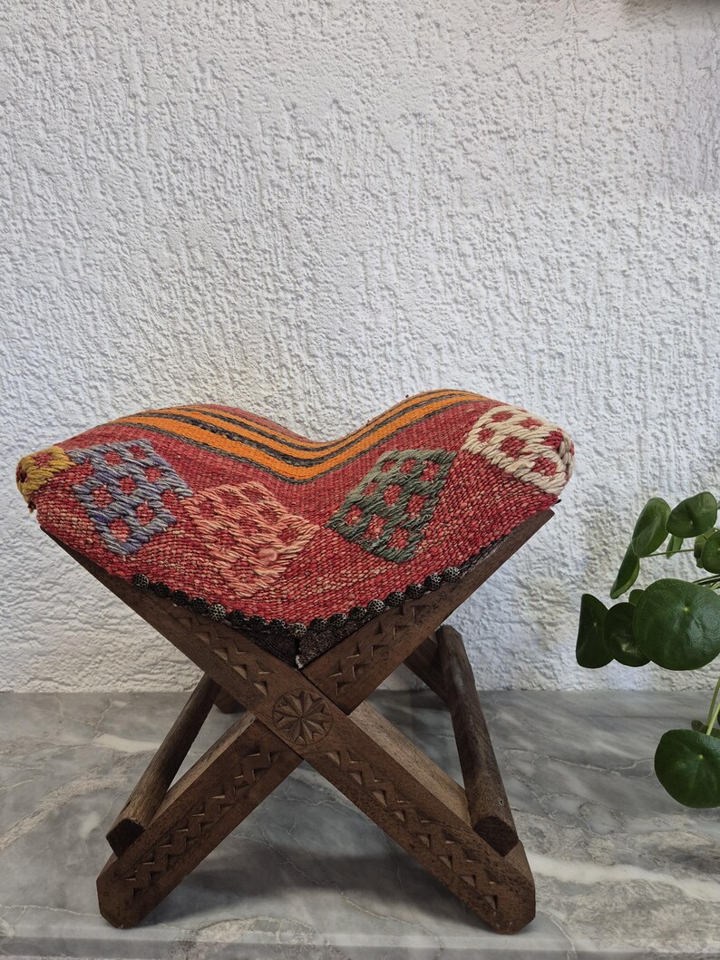 May include: A small, folding wooden stool with a patterned seat. The seat has a red background with colourful geometric designs. The wooden frame has carved details. The stool is designed for sitting and is a decorative item.