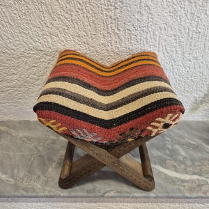 May include: A small wooden stool with a woven seat. The seat has a chevron pattern with red, orange, black, and cream stripes. The wooden legs are carved with floral patterns, creating a rustic and decorative piece, suitable for a home.