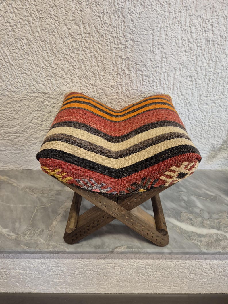 May include: A small wooden stool with a woven seat. The seat has a chevron pattern with red, orange, black, and cream stripes. The wooden legs are carved with floral patterns, creating a rustic and decorative piece, suitable for a home.