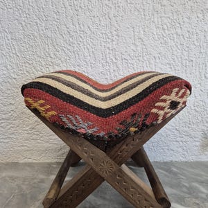 May include: A small, foldable wooden stool with a patterned seat. The seat has a striped design in red, black, beige, and white. The wooden legs are intricately carved with geometric patterns, creating a rustic aesthetic.