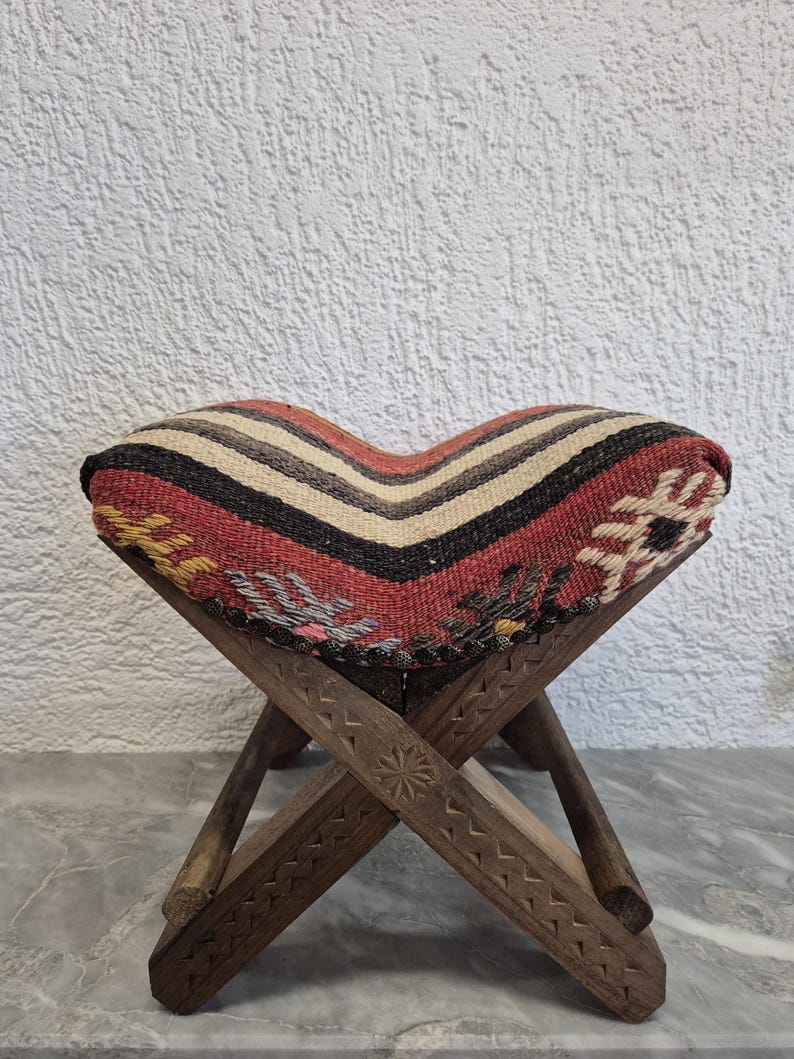 May include: A small, foldable wooden stool with a patterned seat. The seat has a striped design in red, black, beige, and white. The wooden legs are intricately carved with geometric patterns, creating a rustic aesthetic.