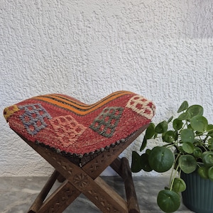 May include: A small, foldable wooden stool with a woven, patterned seat. The seat has a red background with colourful geometric designs. The wooden legs are intricately carved. The stool is approximately 30 cm tall.