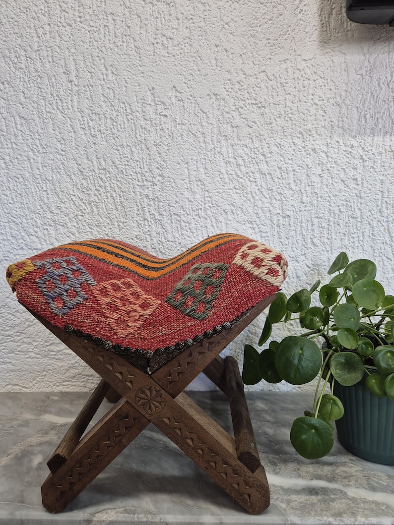 May include: A small, foldable wooden stool with a woven, patterned seat. The seat has a red background with colourful geometric designs. The wooden legs are intricately carved. The stool is approximately 30 cm tall.