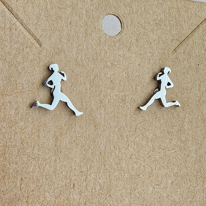Puede incluir: Un par de pendientes plateados con la silueta de una persona corriendo. Cada pendiente muestra una figura en movimiento. Se exhiben sobre una tarjeta marrón con un agujero circular en la parte superior.