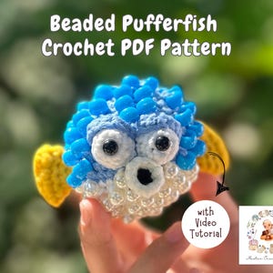 May include: A crocheted blue and white pufferfish with yellow fins and blue beads. The pufferfish is being held in a hand. The image includes the text "Beaded Pufferfish Crochet PDF Pattern" and "with Video Tutorial".
