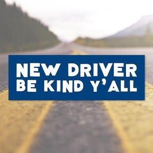Peut inclure: Un panneau rectangulaire bleu foncé avec le texte blanc NEW DRIVER BE KIND Y'ALL. Le panneau est centré sur un arrière-plan flou représentant une route goudronnée avec des marquages jaunes s'étendant au loin sous un ciel lumineux.