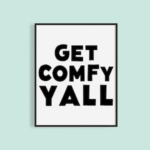 May include: A framed print with the words "GET COMFY YALL" in bold, black letters on a white background. The print is in a black frame and set against a light blue background.