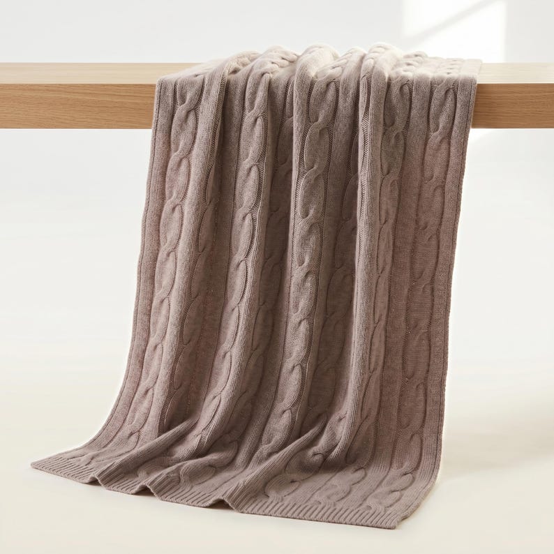 May include: A cable-knit blanket in a muted taupe color is draped over a wooden surface. The blanket's texture is visible, with thick, vertical cable patterns. The soft, neutral color and classic design suggest a cozy home decor item.