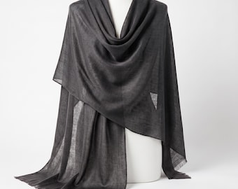 May include: A charcoal gray rectangular scarf draped over a display form. The scarf features a lightweight, semi-sheer woven texture with visible horizontal and vertical threads and delicate fringed edges along the bottom hemline.