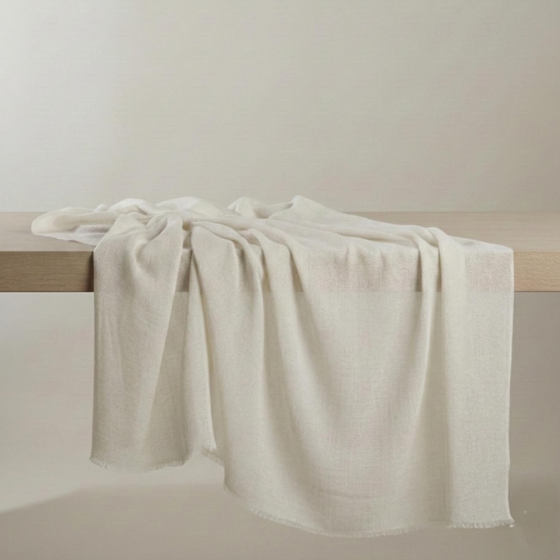 May include: A soft, off-white rectangular throw blanket with a fine, lightweight knit texture and delicate frayed edges. The fabric drapes naturally, showcasing a neutral, solid color and a consistent, semi-sheer weave throughout the entire piece.