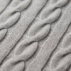 May include: Close-up of a gray cable knit fabric. The knit pattern features thick, intertwined strands creating a textured surface. The color is a solid, neutral gray, suitable for various apparel items.