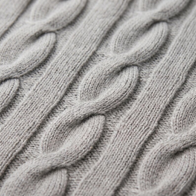 May include: Close-up of a gray cable knit fabric. The knit pattern features thick, intertwined strands creating a textured surface. The color is a solid, neutral gray, suitable for various apparel items.