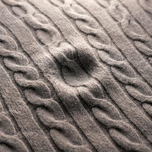 May include: Close-up of a gray cable knit fabric. The knit pattern features vertical rows of intertwined cable designs, creating a textured surface. The fabric appears soft and warm, suitable for sweaters or blankets.