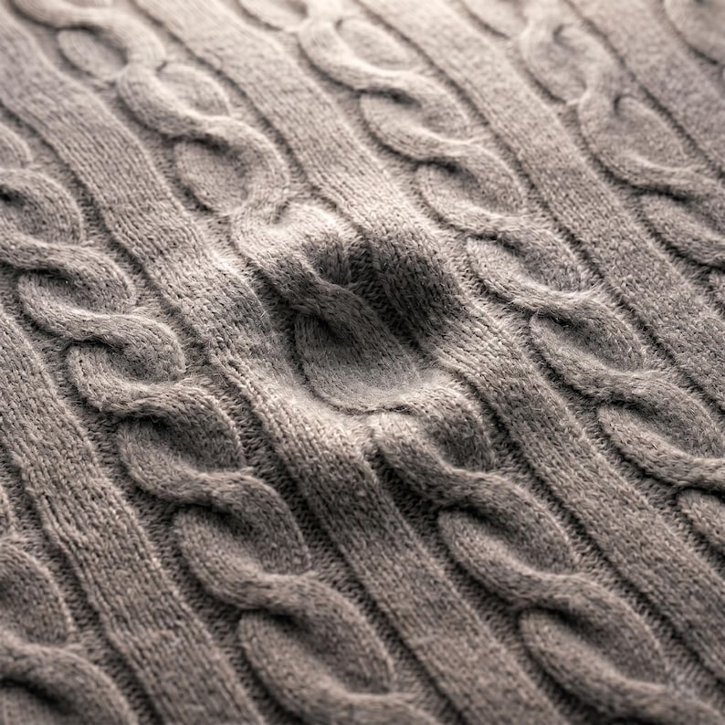 May include: Close-up of a gray cable knit fabric. The knit pattern features vertical rows of intertwined cable designs, creating a textured surface. The fabric appears soft and warm, suitable for sweaters or blankets.