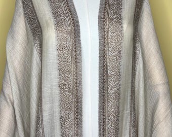 May include: A beige open-front shawl featuring a dark brown intricate floral and paisley border pattern along the front edges and hem. The fabric has a subtle vertical texture and drapes over the shoulders, showcasing a symmetrical design.
