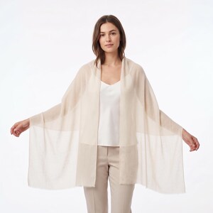 May include: A lightweight, rectangular beige shawl draped over the shoulders. The garment features a fine, semi-sheer knit texture with clean, straight edges, providing a soft and neutral appearance that complements the layered outfit underneath.