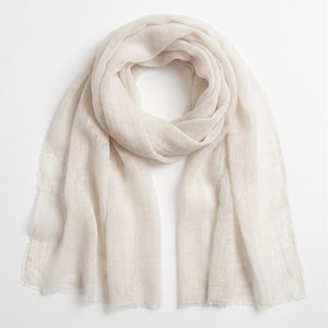 May include: A lightweight, semi-sheer scarf in a soft off-white or cream color. The fabric features a fine, loose weave with a subtle texture and delicate frayed edges along the ends, creating a soft, airy appearance for this rectangular accessory.