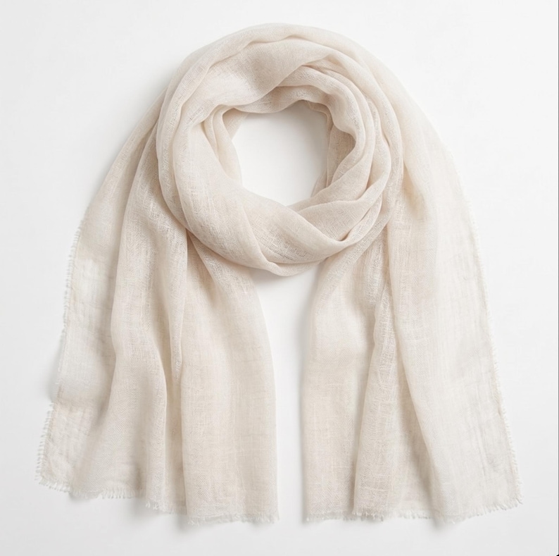 May include: A lightweight, semi-sheer scarf in a soft off-white or cream color. The fabric features a fine, loose weave with a subtle texture and delicate frayed edges along the ends, creating a soft, airy appearance for this rectangular accessory.