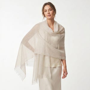May include: A lightweight, semi-sheer beige shawl with a fine, woven texture and delicate fringe detailing along the edges. The fabric drapes loosely over the shoulders, showcasing a neutral, solid color and a soft, airy appearance throughout.