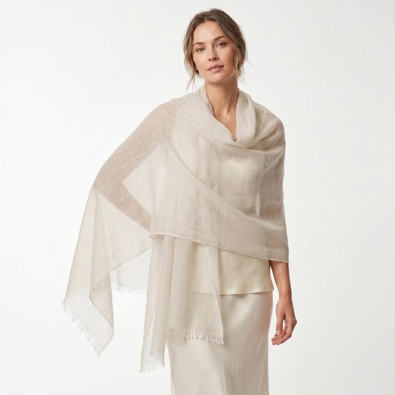 May include: A lightweight, semi-sheer beige shawl with a fine, woven texture and delicate fringe detailing along the edges. The fabric drapes loosely over the shoulders, showcasing a neutral, solid color and a soft, airy appearance throughout.