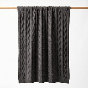 May include: A dark gray cable-knit blanket is draped over a wooden rod against a white background. The blanket's texture is visible, with vertical cable patterns running its length. The blanket appears soft and cozy, suitable for home decor.