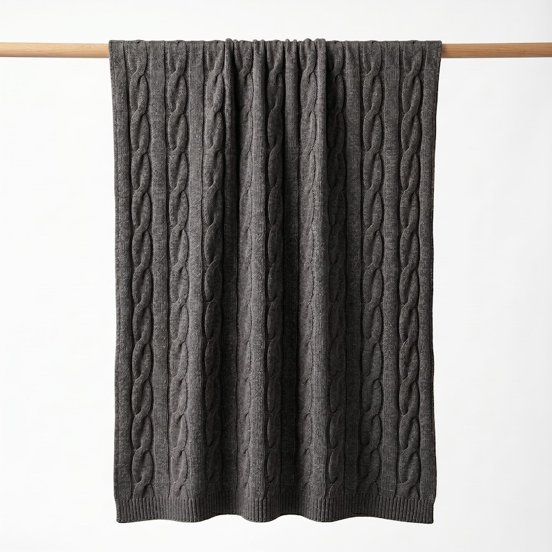 May include: A dark gray cable-knit blanket is draped over a wooden rod against a white background. The blanket's texture is visible, with vertical cable patterns running its length. The blanket appears soft and cozy, suitable for home decor.