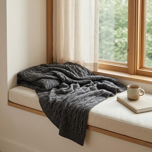 May include: A dark gray cable-knit throw blanket draped over a window seat. The blanket is folded and has a textured pattern. A small mug with a handle and a book are also on the seat.