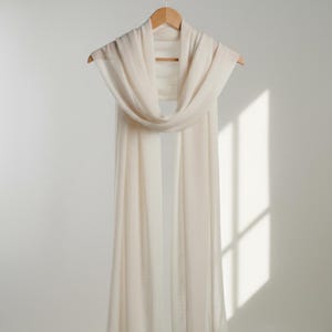 May include: A long, rectangular scarf in a solid off-white color, draped over a wooden hanger. The fabric appears lightweight and semi-sheer with a fine, soft texture, hanging in vertical folds that extend downward from the hanger's horizontal bar.