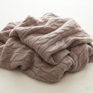 May include: A folded, taupe-colored cable knit blanket. The blanket has a textured pattern of interwoven cables and a ribbed edge. The soft, neutral color and thick knit suggest warmth and comfort. The blanket is made of a soft material.