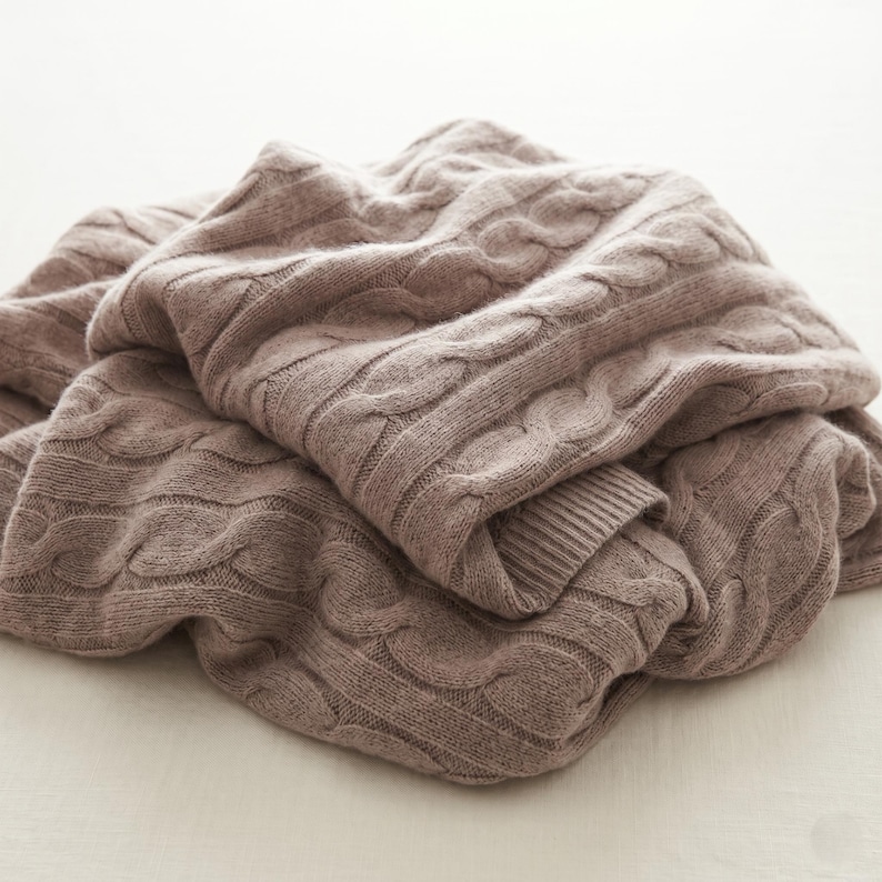 May include: A folded, taupe-colored cable knit blanket. The blanket has a textured pattern of interwoven cables and a ribbed edge. The soft, neutral color and thick knit suggest warmth and comfort. The blanket is made of a soft material.
