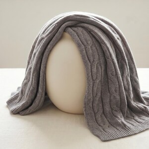 May include: A gray cable-knit blanket draped over a white, rounded form. The blanket has a textured pattern and a soft appearance. The neutral color palette and simple design suggest a focus on comfort and warmth, suitable for home decor.
