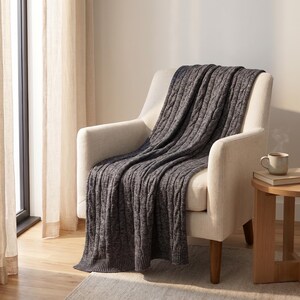 May include: A gray cable-knit throw blanket draped over a cream-colored armchair. The blanket has a textured pattern and is made of a soft material. A small wooden side table holds a mug and a stack of books.