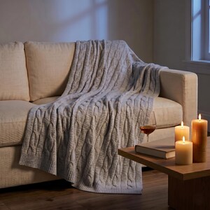 May include: A light gray cable-knit throw blanket draped over a beige sofa. The blanket is thick and textured. A glass of wine, a book, and lit candles sit on a wooden side table. The scene is lit by soft, warm light.