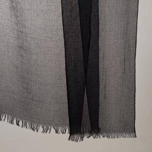 K&ouml;nnte beinhalten: Ein leichter Schal mit einem halbtransparenten, strukturierten Gewebe in Anthrazit und Schwarz. Der Stoff weist ein dezentes, gitterartiges Muster auf, und die Unterkante ist mit feinen, losen Fransen versehen, die das Design abrunden.