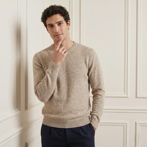 Könnte beinhalten: Ein hellbrauner Rundhals-Pullover. Der Pullover ist aus einem weichen, strukturierten Material gefertigt. Die Person auf dem Bild trägt den Pullover mit einer dunkelblauen Hose. Der Pullover hat lange Ärmel und ein klassisches, schlichtes Design.