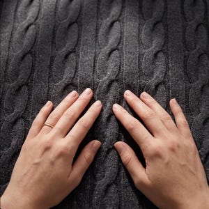 May include: Close-up of a dark gray cable-knit sweater. The textured pattern of the knitwear is visible, with a series of raised, intertwined cable designs. The sweater's color is a solid, neutral gray.